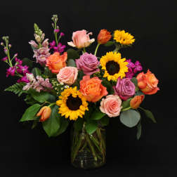 Mixed bouquet of roses, sunflowers, and purple blooms in a glass vase