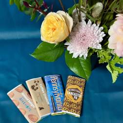 Floral arrangement with pink and yellow blooms beside four wrapped chocolate bars on teal fabric.