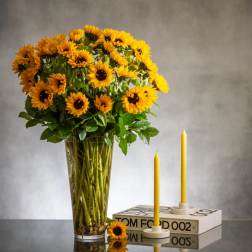 Tall arrangement of yellow sunflowers in a clear vase beside two yellow taper candles on a book.