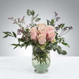 Arrangement of pale pink roses with small purple blooms in a clear glass vase.