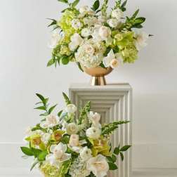 Two white and green floral arrangements in gold pedestal vases