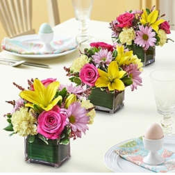 Three small floral centerpieces in square glass vases on a table