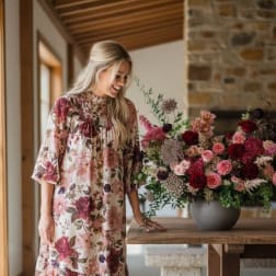 Large pink and burgundy flower arrangement in a gray bowl on a wooden table beside a smiling woman.
