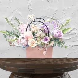 Pastel roses and lavender flowers arranged in a pink gift bag with ribbon handles.