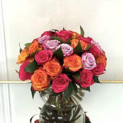 Round bouquet of hot pink, orange, and lavender roses arranged in a clear glass vase.