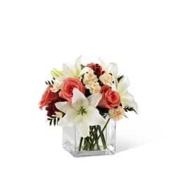 Low arrangement of white lilies, peach roses, and cream carnations in a clear glass cube vase