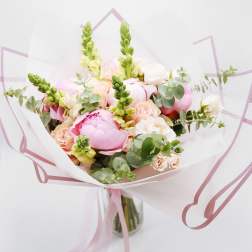Hand-tied bouquet of pink peonies, peach roses, and snapdragons wrapped in white paper in a glass vase
