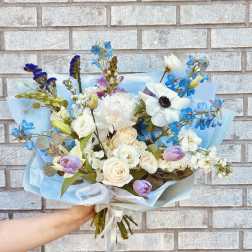 Handheld bouquet of white, blue, and lavender flowers wrapped in pale blue paper