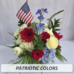 Patriotic red, white, and blue flower arrangement with roses, gerbera daisies, hydrangeas, delphinium, and a small American flag.