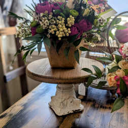 Basket-style arrangement of purple roses and white blooms with green accents in a brown container.