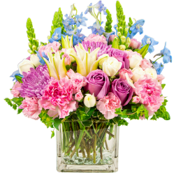 Mixed pink, purple, and white flower arrangement with lilies and roses in a clear square glass vase