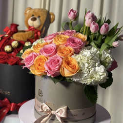 Pink and orange roses with white hydrangeas in a round box, with a teddy bear behind it.