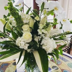 Tall arrangement of white lilies, roses, orchids, and hydrangeas with bold green leaves in a clear glass vase