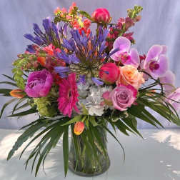 Bright mixed arrangement of pink gerberas, roses, orchids, and other flowers in a clear glass vase