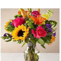 Colorful mixed bouquet in a clear glass vase with sunflowers, roses, lilies, and purple blooms