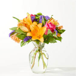 Bright mixed bouquet of lilies, roses, and purple filler flowers in a clear glass vase