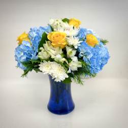 Yellow roses, white blooms, and blue hydrangeas arranged in a blue glass vase