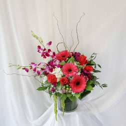 Bouquet of pink gerbera daisies, red roses, and purple orchids in a glass vase