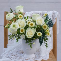White roses and daisy-like flowers arranged in a white vase with lace draping.