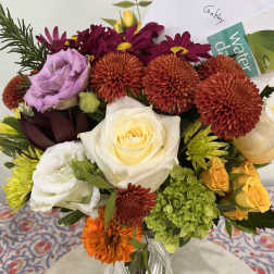 Mixed bouquet of orange mums, white roses, purple lisianthus, and green hydrangea in a glass vase with a card.