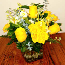 Yellow roses arranged in a square glass vase with greenery.