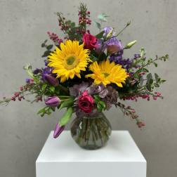 Bouquet of yellow gerbera daisies, pink roses, and purple flowers in a glass vase