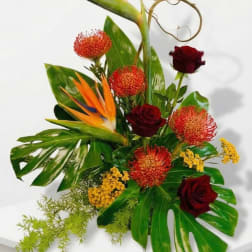 Tall tropical arrangement with bird of paradise, red roses, orange protea and yellow blooms on large glossy leaves