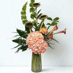 Tall floral arrangement with pink hydrangea, sunflowers, calla lilies, and a glass vase