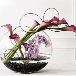 Calla lilies and purple orchids in a round glass bowl with black stones