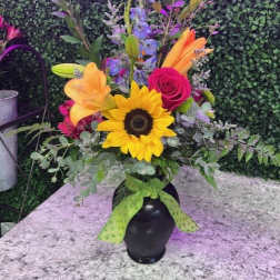 Tall mixed bouquet with sunflower, lilies, rose, and blue delphinium in a black vase with green bow.
