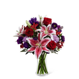 Mixed bouquet of pink lilies, red roses, and purple flowers in a clear glass vase