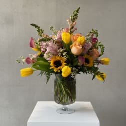 Mixed bouquet of tulips, roses, sunflowers, and pink blooms in a glass vase
