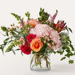 Mixed bouquet of pink, peach, and red flowers in a clear glass vase