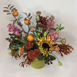 Autumn floral arrangement with sunflowers, roses, pink blooms, and oranges in a green vase