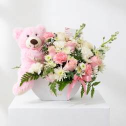 Pink teddy bear beside a pastel flower arrangement in a white box