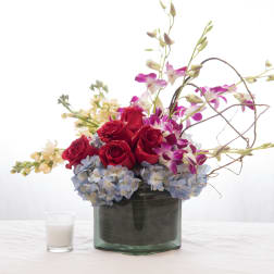 Low arrangement of red roses, purple orchids, and blue hydrangeas in a glass vase with a small white candle.