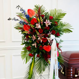 Tall funeral spray with red roses, lilies, and tropical leaves beside a casket