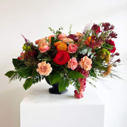 Mixed bouquet with red roses, peach carnations, and orange blooms in a black vase