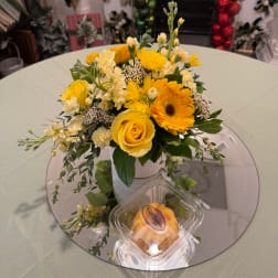 Yellow and white flower arrangement in a textured vase with a pastry container in front
