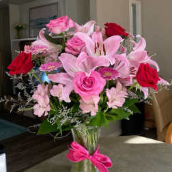 Pink lilies and roses arranged in a clear glass vase with a pink ribbon.