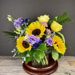 Mixed bouquet with sunflowers, yellow roses, and purple blooms in a glass vase