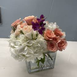 Pink roses and white hydrangeas in a clear square vase with purple orchids