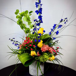 Tall mixed floral arrangement with blue delphinium, yellow roses, and pink blooms in a patterned vase