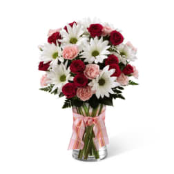 Tall bouquet of red roses, white daisies, and pink carnations in a clear glass vase with a pink ribbon.