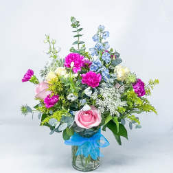 Mixed bouquet of pink, blue, and white flowers in a glass vase with a blue ribbon