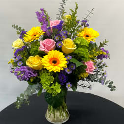 Mixed bouquet of yellow gerbera daisies, pink and yellow roses, and purple flowers in a glass vase