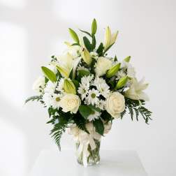 White lilies, roses, and daisies arranged in a glass vase with a ribbon