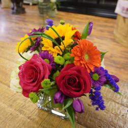 Mixed bouquet with roses, sunflowers, and gerbera daisies in a square glass vase