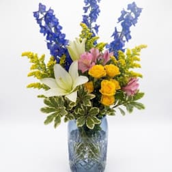 Blue and yellow mixed bouquet in a glass vase with white lilies and pink flowers