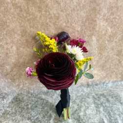 Small boutonniere with dark burgundy ranunculus, yellow and white accents, wrapped with black ribbon